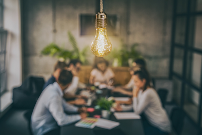 Innovation entsteht im Team – neue Ideen gemeinsam entwickeln Leuchtende Glühbirne als Symbol für Innovation und Ideen, im Hintergrund ein Team bei einer Besprechung